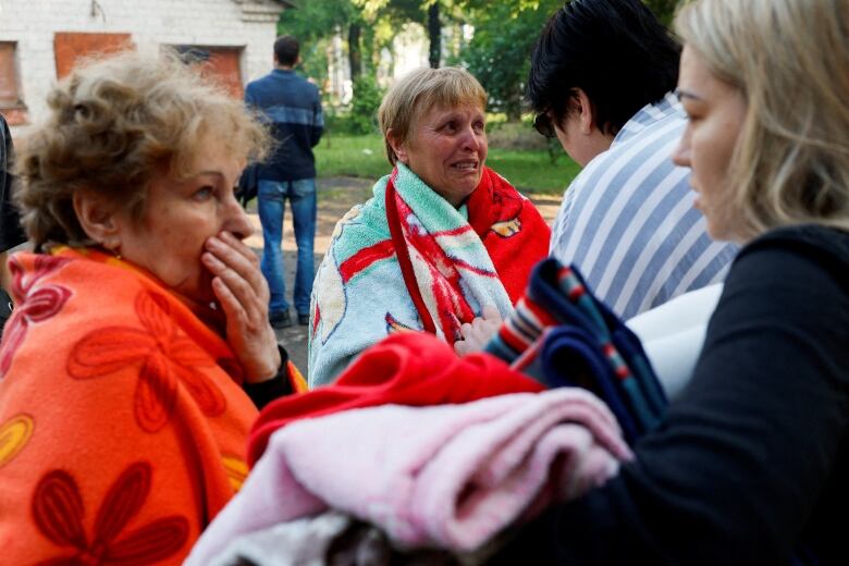 Two women appear in shock as they are wrapped in bath towels. The woman on the left, wrapped in an orange towel, holds her hand to her face.