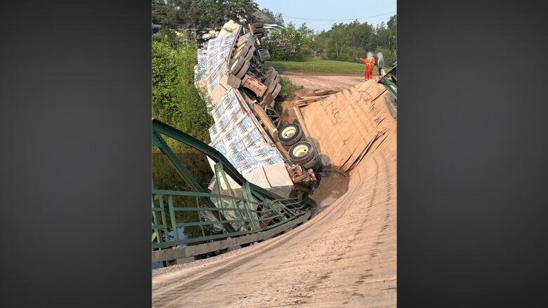 A transport truck is shown flipped on its side in a river next to a collapsed bridge.