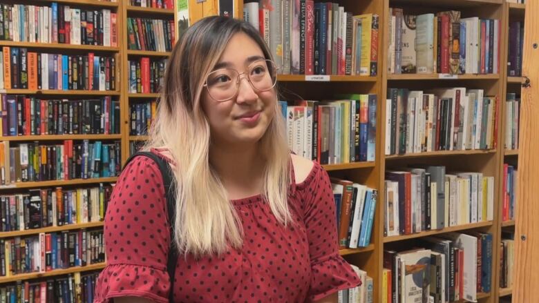 A woman in a red shirt and glasses is pictured standing in a bookstore. 