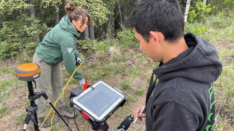 A woman in a green jacket and a man in a black hoodie adjust a screen on a piece of radar machinery while standing in a patch of grass near a wooded area.