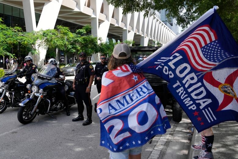 A person with their back to the camera wearing a ballcap and waving a flag is shown standing near police officers.