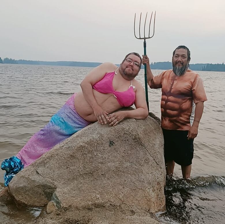 A man lies on a rock in a merman costume with a colorful fin and pink bra, while a man dressed in a shirt with a pattern showing rippled abdominal muscles stands beside him, holding a pitchfork. 