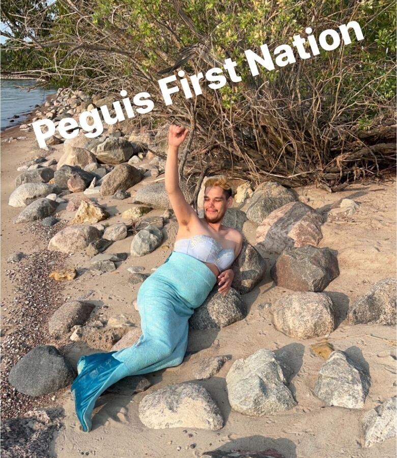 A man lies on a sandy beach in a merman costume with a bright blue fin and light coloured bra.