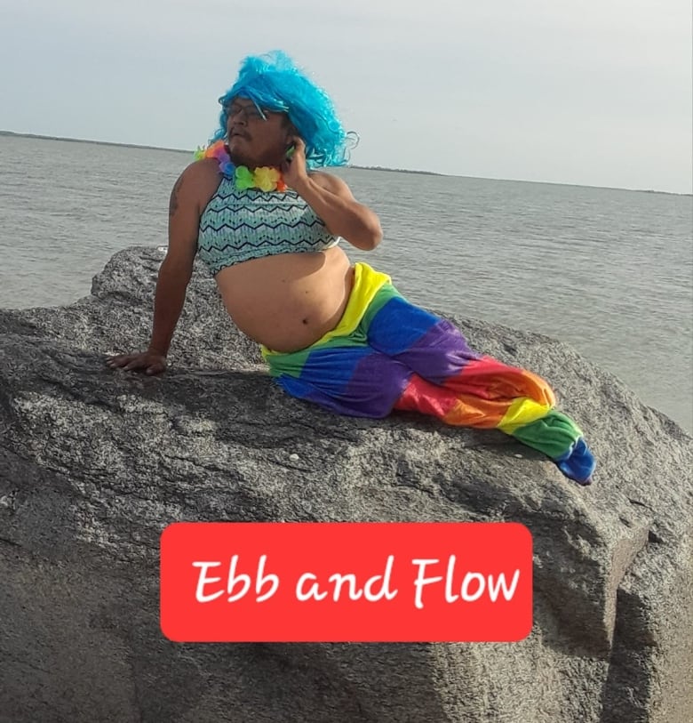 A man lies on a rock in a merman costume with a rainbow coloured fin, patterned bra and blue hair.