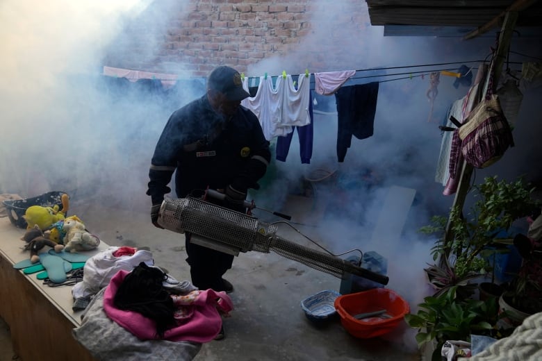 A man in a blue jacket, pants and cap stands in a yard, with clothes hanging on a line behind him, as he holds a long metal machine spraying fog. 