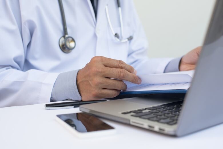 Doctor reading patient health record chart and working on laptop computer on white desk in hospital with copy space, electronic health records system EHRs, teleconference or telemedicine concept.