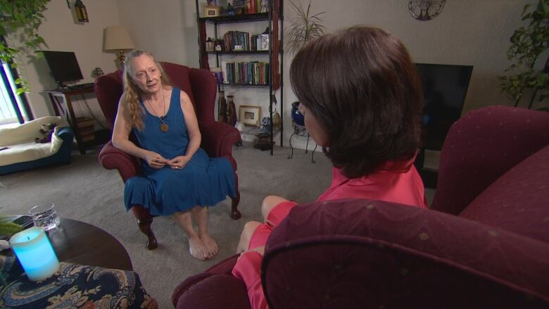 A woman, Constance McCall, is wearing a sleeveless blue mid-calf length dress sits in an upholstered chair with her hands in her lap, speaking to a brown-haired woman, Rosa Marchitelli, whose back is to the camera. McCall's long hair is pulled back from her face. Both women are seated in a living room with grey carpet and a bookshelf and computer monitor in the background.