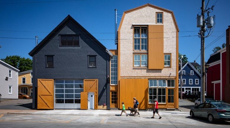 People walk by a black and orange pair of wooden buildings, one two story with a peaked roof and the other higher.