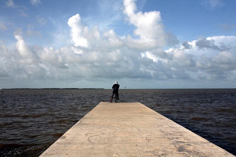 A man stands at the end of a long cement pier, looking out at the ocean.