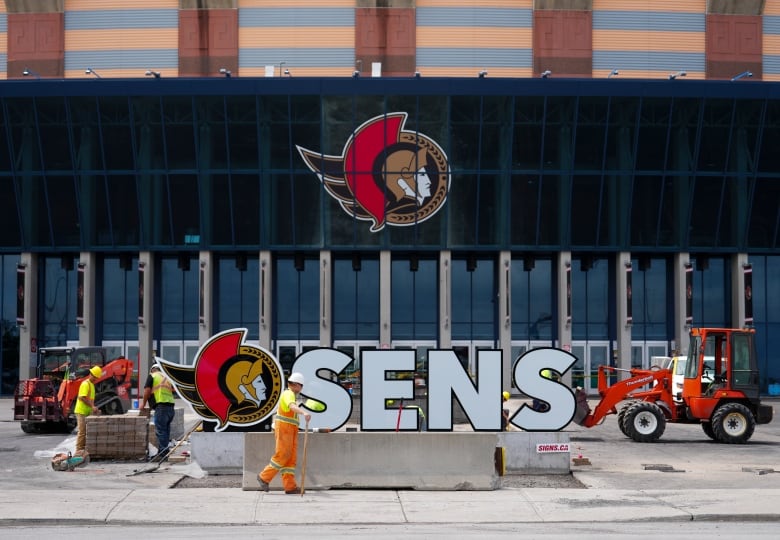 Landscapers around the Ottawa Senators Roman general-style logo on an arena with a large white S-E-N-S sign on the ground for photo ops.