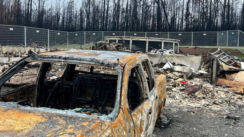 A burnt-out car is shown in front of a burnt property surrounded by white fencing. 