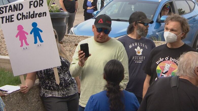 A man in shades stands with a cellphone, with a sign behind him reading, 