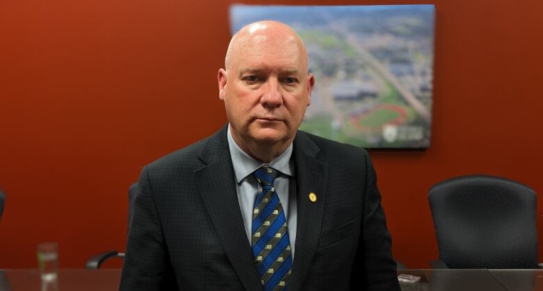 Man with serious expression stands in an office, wearing a business suit.