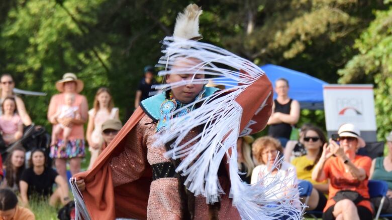 Woman in traditional Indigenous clothing dances in front of a crowd on grassy area