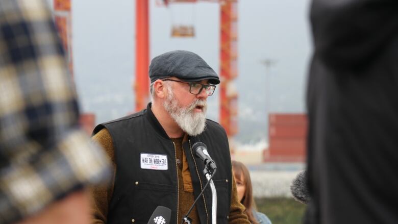 A man with a black hat, glasses, and grey beard speaks into a microphone on a beach along Vancouver's downtown waterfront