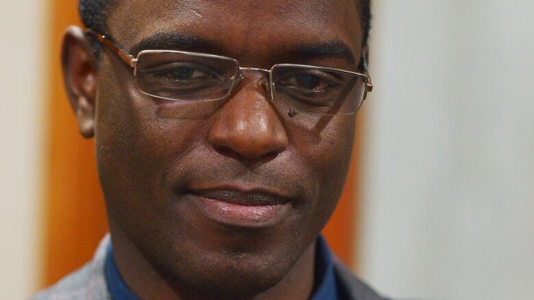 A man is pictured in relative close-up. He wears glasses and looks down pensively. He is wearing a grey suit and dark blue shirt.  