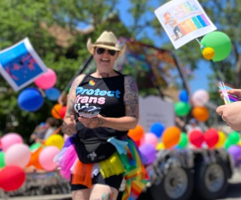 A person wearing a black tank top reading 'Protect trans' and a rainbow-coloured tutu walks in a parade, handing out pamphlets with the transgender colours of baby blue, light pink and white. Behind them is a float covered in multi-coloured balloons.