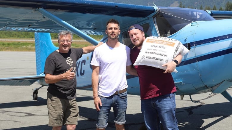 Three men, one of them holding a sack of malt, smile in front of a small blue plane on an airstrip.