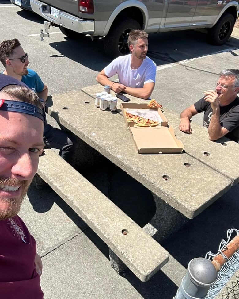 A man takes a selfie of a few men eating pizza on a parking lot bench.