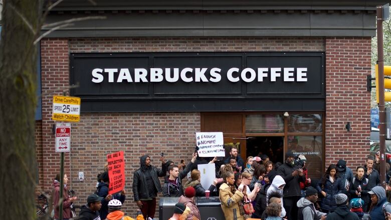 Dozens of people, some holding up signs, are shown outside a brick building housing a Starbucks coffee store.