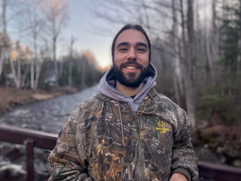 A man in a camouflage sweater smiles. His dark brown hair is in a ponytail and he has a beard. He stands outside in a forest, near a brook.