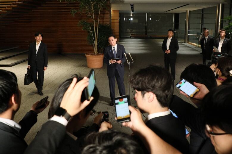 A man in a suit is seen in front of a microphone, surrounded by several people holding up cellphones.