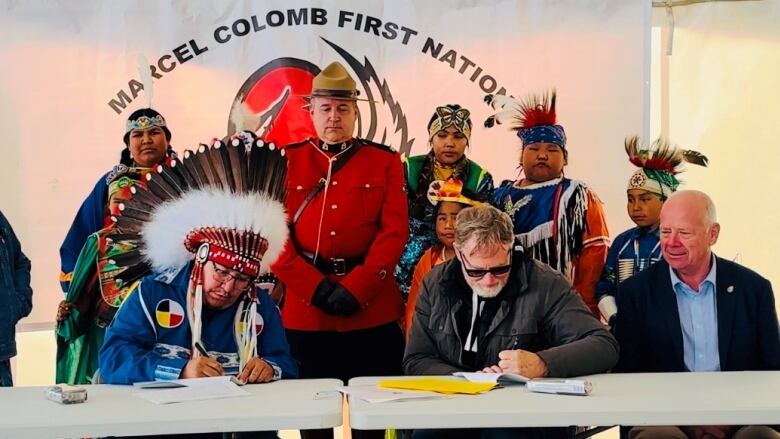 A First Nations chief in traditional clothing and headdress sits at a table alongside businessmen while an RCMP officer in traditional red serge stands behind them.