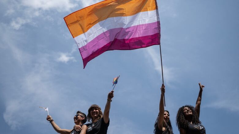 Four people wearing black t-shirts standing outside, with the blue sky in the background. A person, second from right, holds up a large flag striped with the shades of orange, white, pink and rose. 