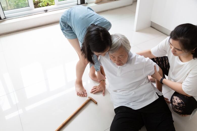 An older woman sits on the ground, leaning back, with a walking stick lying next to her. Two younger women try to help her to her feet. 