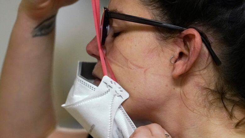 Shot in profile, the image shows a nurse with dark hair and glasses pulling on a while mask. 