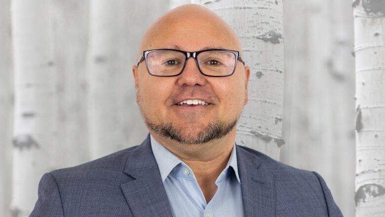 A man in a dark blue blazer and light blue button-up smiles. He wears glasses.