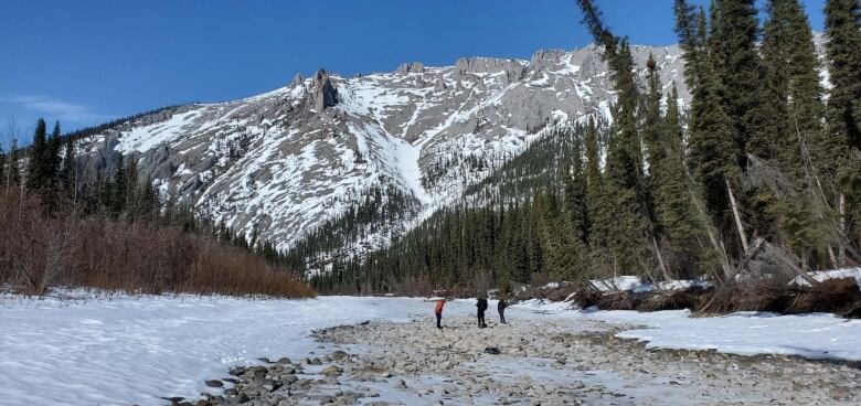 Three people are seen from afar standing in a snowy river bed, with mountains in the background.