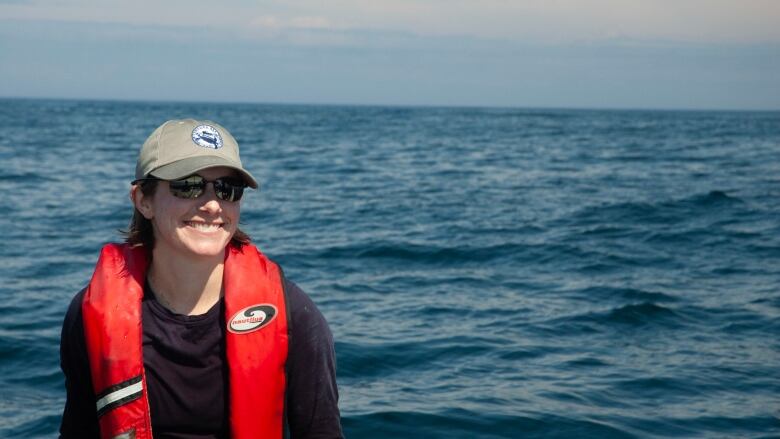 A young woman with a beige baseball hat and dark sunglasses smiles at the camera. She is standing outdoors in front of a body of water wearing a dark long-sleeve shirt and a red-orange life jacket.