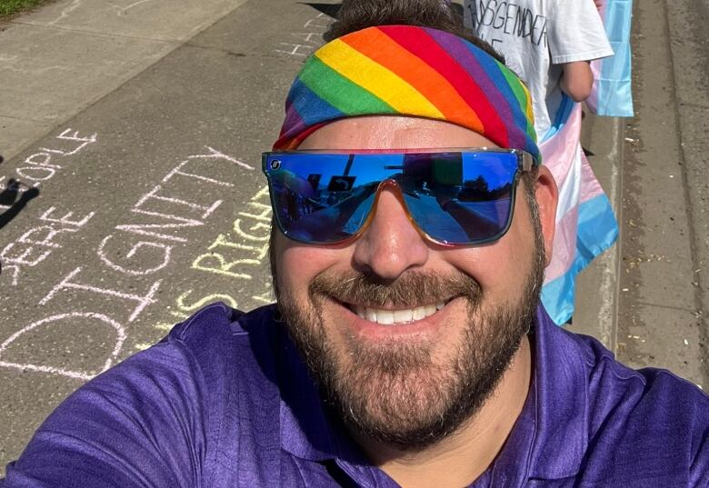 A person stand outside on a sidewalk. They are wearing sunglasses and a rainbow headband.