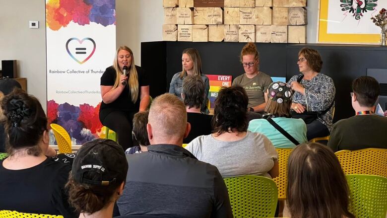 Four women sit on a panel, addressing a crowd in a community space.