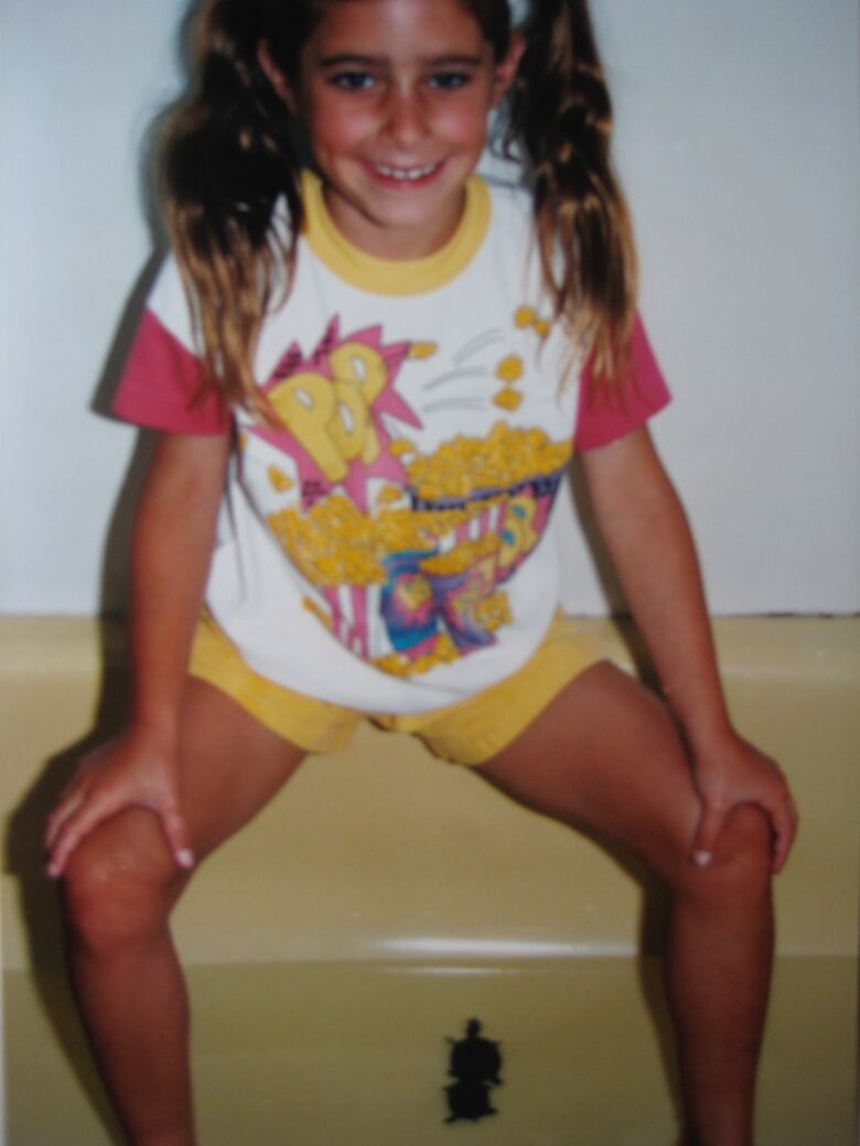 A young girl with long brown hair in pig-tails, wearing a tee-shirt and shorts sits on the edge of a yellow bathtub, with two tiny turtles in the water of the tub below her.