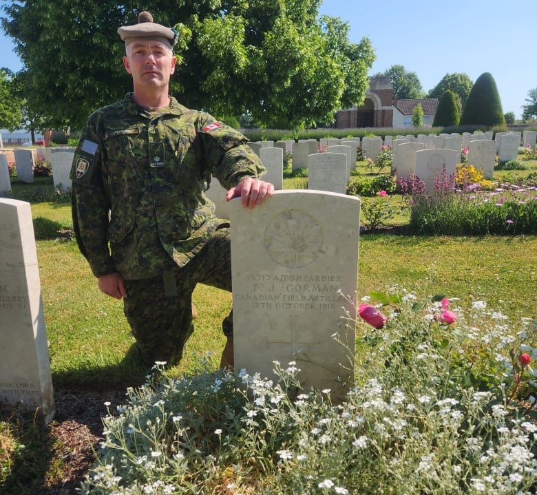 A solider kneels in uniform beside a grave stone in a graveyard with many flowers.