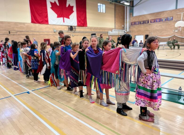 A line of children with their hands on each other's shoulders stand in a gym.