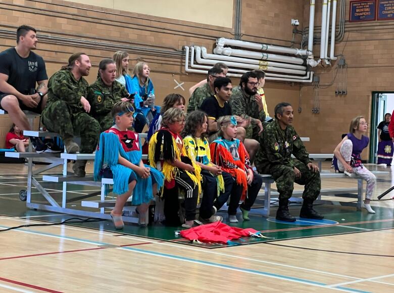 A group of children and military members sit on some bleachers in a gym.