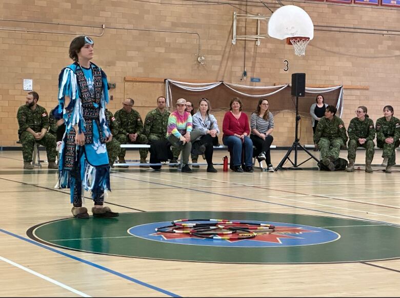 A dancer dressed in blue stands in a gym as people watch from the sidelines.