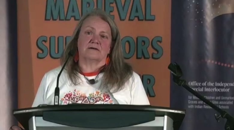 A woman with grey hair is standing at a podium with a microphone. She is wearing a white t-shirt with an Indigenous language on the chest in a colourful array. Behind her is an orange sign with blue letters.