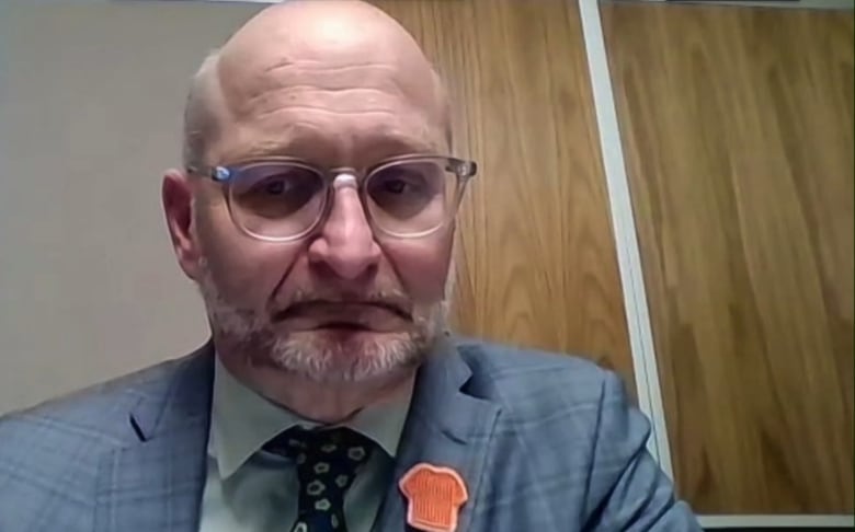 A bald white man with a grey beard is wearing glasses and a suit and tie. He is sitting in an office.