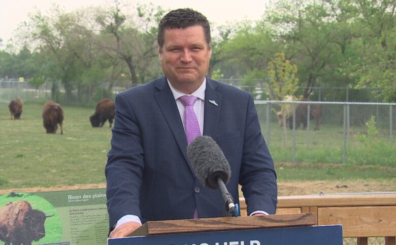 A man stands at a podium, in front of an enclosure of bisons.