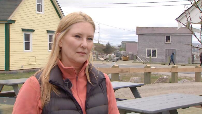 A white woman with long blond hair wears a black puffy vest over a pink sweater. She is standing among picnic tables outside with colourful homes and a winding road in the background.