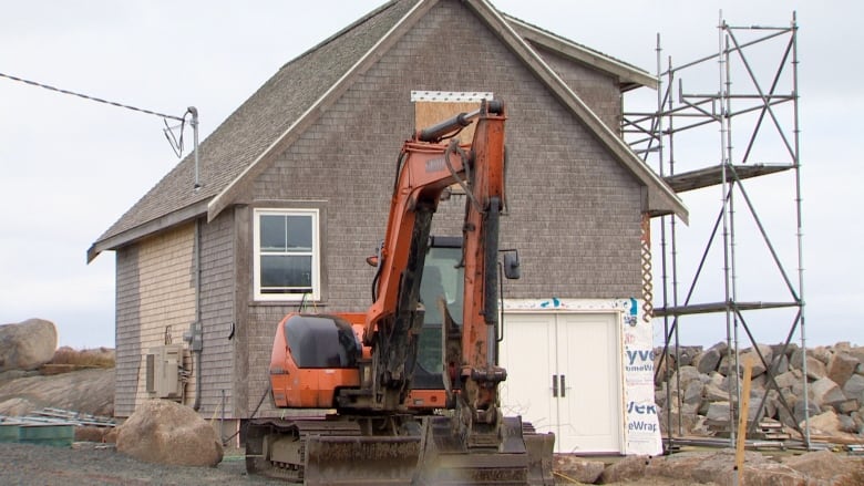 An orange front-end loader stands outside a grey shingled wooden barn that is clearly under construction