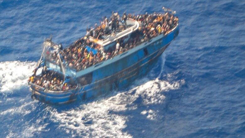 A blurry image shows dozens of people on the deck of a battered blue fishing boat in the Mediterranean Sea. 