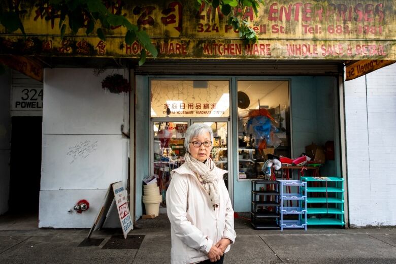 A woman in white jacket stands in front of a shop.