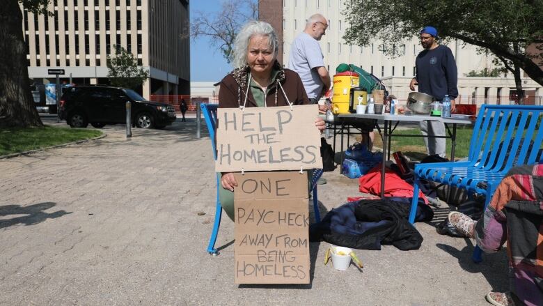 Woman sitting down with two signs. The top sign says help the homeless. The bottom sign says one paycheck from being homeless. She is sitting outside of Regina City Hall. People are cooking in the background, 