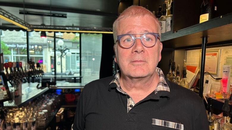 A man wearing a black polo shirt and grey glasses stands in front of a bar. 