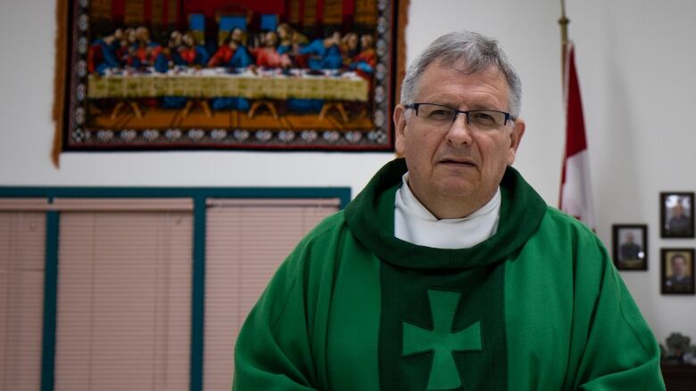 A white man with short grey hair and glasses, is wearing a green cloak with a cross on the chest. He is standing in a church office. A painting of the last supper is hung on the hall behind him.